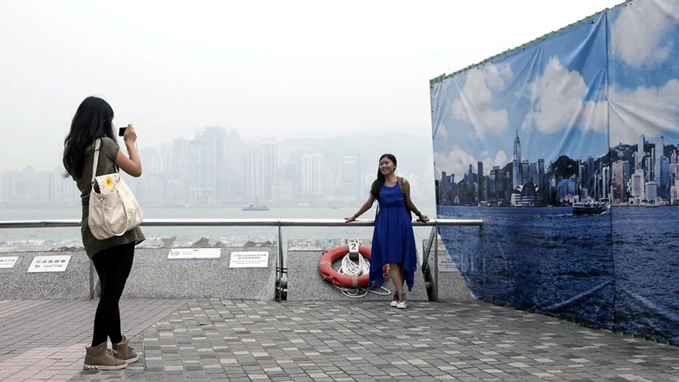 Turisták félnyképezkednek egy tiszta levegőjű Hongkong látképét ábrázoló transzparens előtt Hongkongban, szmog