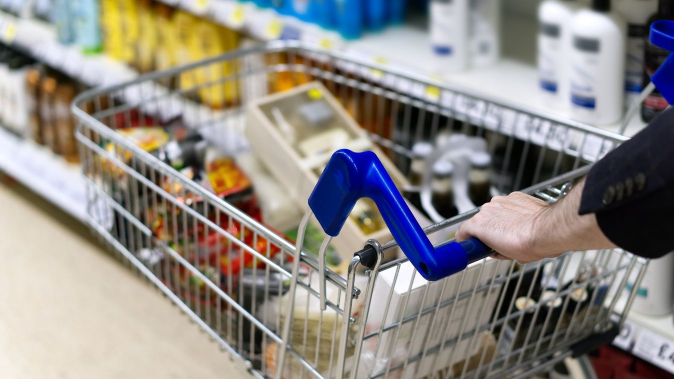 A man's arms pushing a shopping trolley full of groceries along a supermarket aisle. 
