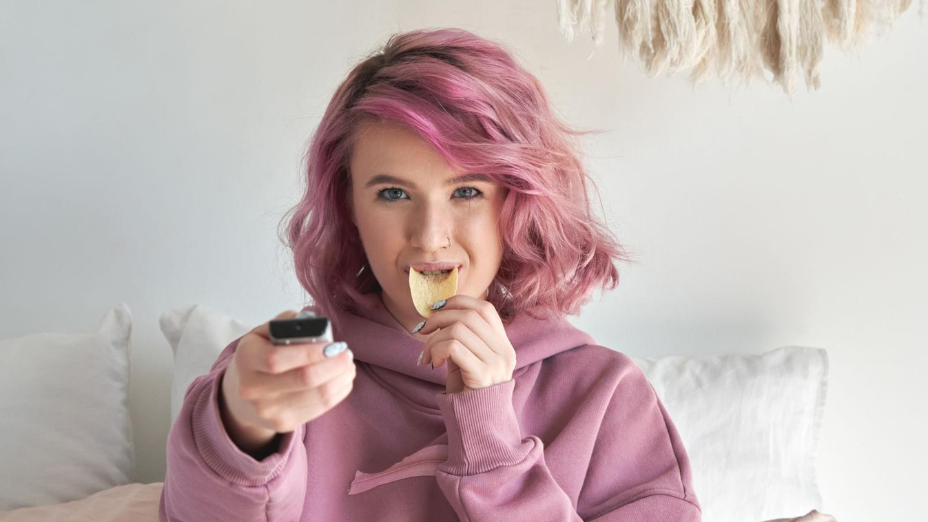 Happy hipster gen z teen girl with pink hair watching tv eating chips in bed. Happy hipster gen z teen girl with pink hair watching tv movie series sitting in bed, holding remote control, eating chips snack in bed. Teenager relaxing watching television re