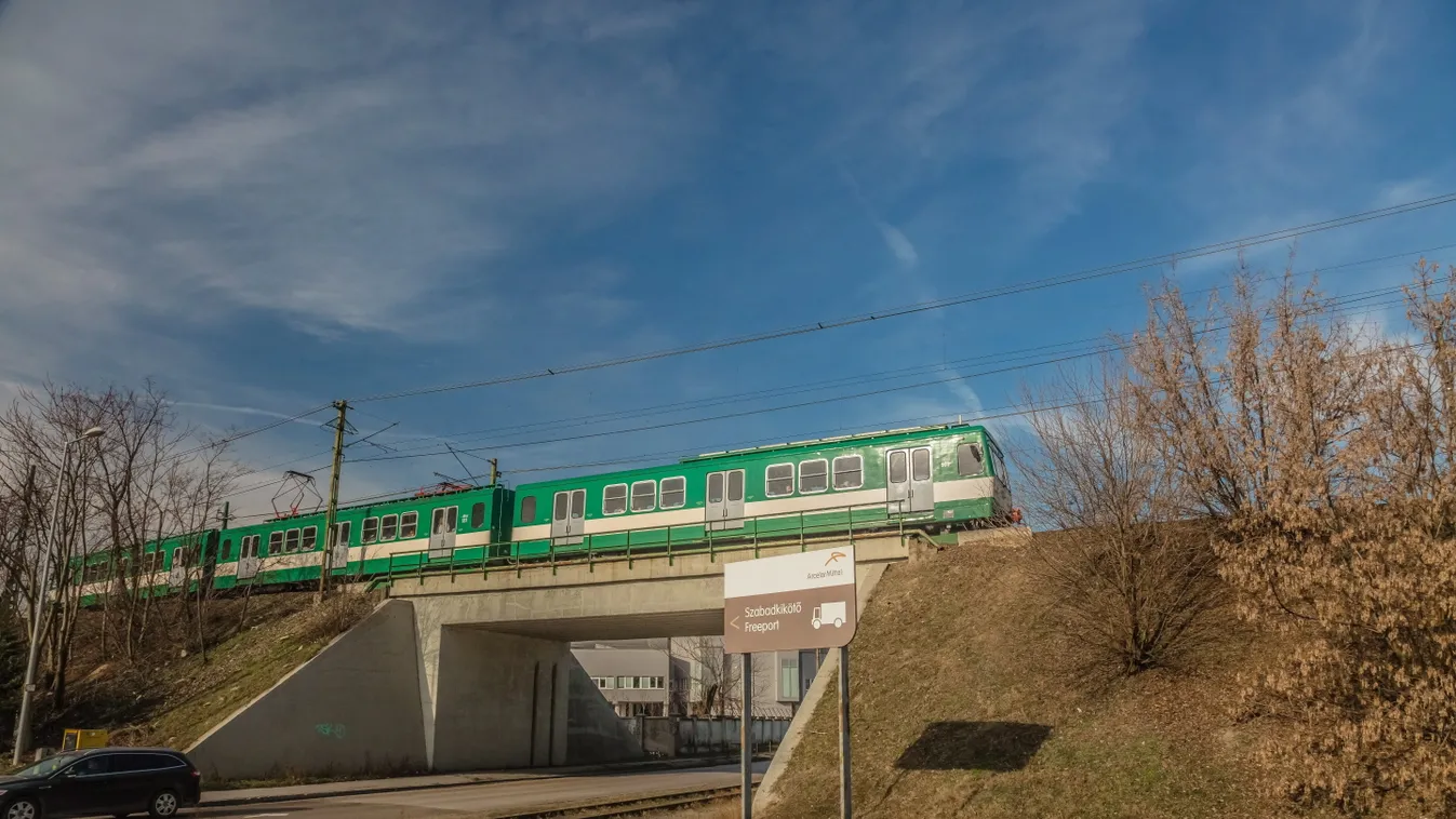 aluljáró BKK-BKV H7-es HÉV csepeli Hév ég fa felhő felirat felsővezeték kopasz fa KÖZLEKEDÉSI ESZKÖZ KÖZLEKEDÉSI LÉTESÍTMÉNY KÖZTERÜLET NÖVÉNY személyautó szerelvény tájékoztató tábla vasúti töltés villanyoszlop 