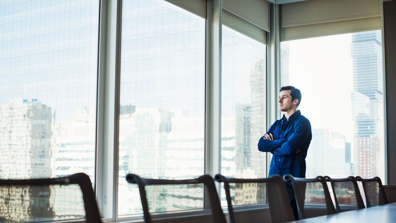 A man standing with arms crossed in a meeting room looking out of a window at an urban landscape. STANDING Working 25-30 Years Male Men Young Men Young Adult Man City Life 1 Person USA New York New York City Colour Image Photography Camera Focus Selective