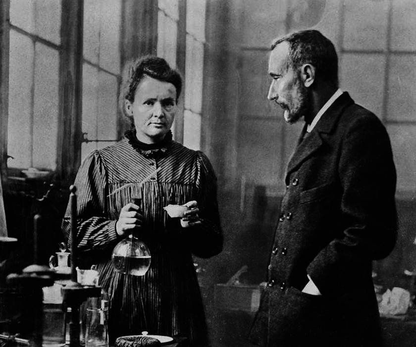 Marie Curie kezében lombikot tart, mellette férje Pierre Curie