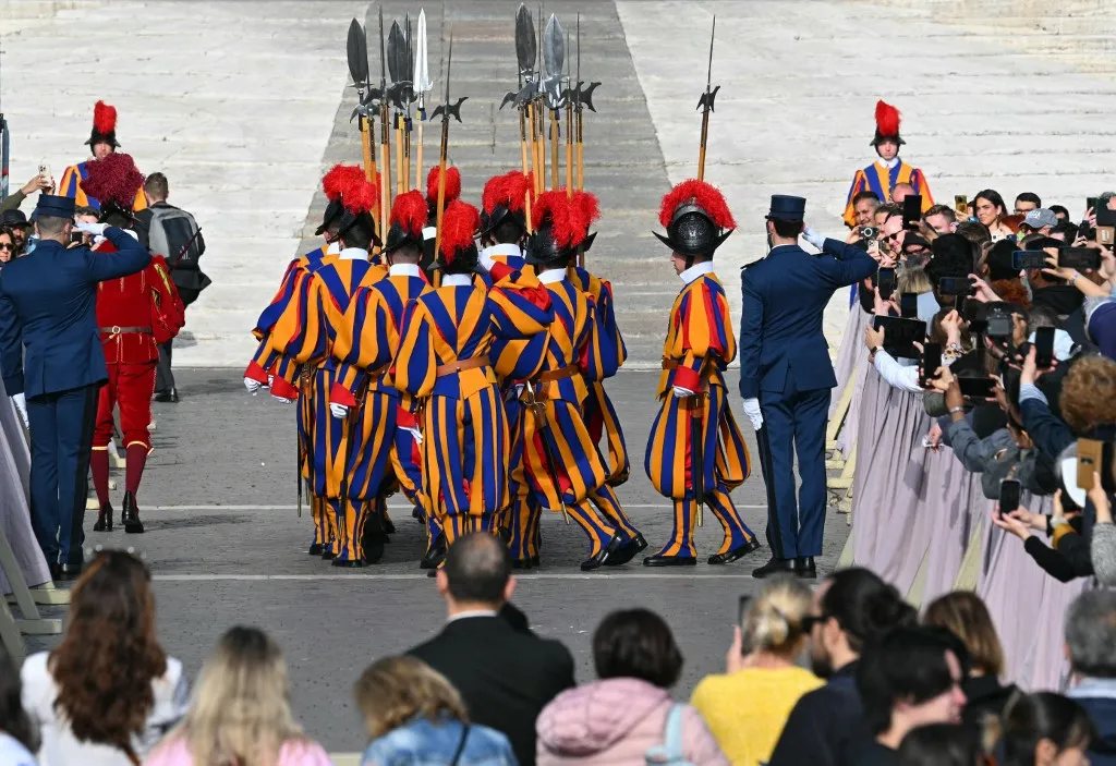 Swiss guards march next to attendees at St Peter's Square prior to the arrival of the coffin of the late Pope, set to be transported from the chapel of Santa Marta to St Peter's Basilica, following the Pope's death, in the Vatican on April 23, 2025. The Pope died of a stroke, the Vatican announced hours after the death on April 21, 2025, of the 88-year-old reformer who inspired devotion but riled traditionalists during 12 years leading the Catholic Church. (Photo by Andreas SOLARO / AFP)