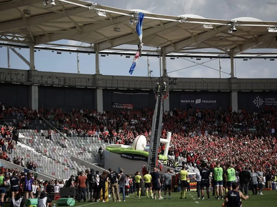 Spectators watch as firefighters work to rescue an air force parachutist (Top) whose canopy is jammed on the roof of the stadium prior to the European Rugby Champions Cup last 16 rugby union match between Stade Toulousain Rugby (Toulouse) and Sale Sharks at the Stadium in Toulouse, south-western France, on April 6, 2025. An air force parachutist, who was supposed to bring the match ball onto the pitch at Toulouse Stadium from the air, had to be unhooked from the roof of the stadium before the start of the Champions Cup Round of 16 match, which was delayed on April 6, 2025. Five minutes before kick-off of the match against England's Sale, scheduled for 4 p.m., a gust of wind knocked the parachutist off course, sending him towards the roof where his canopy became caught in the stadium structure. (Photo by Valentine CHAPUIS / AFP), ejtőernyős