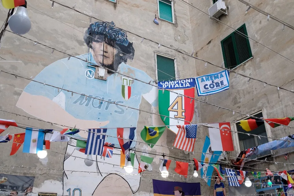 A flag depicting the Scudetto with the number four and the inscription "Campioni d'Italia" (Champions of Italy) is seen near a mural of Diego Armando Maradona in the Quartieri Spagnoli, downtown Naples, on May 24, 2025 as Napoli won the fourth Italian football Champions after the match with Cagliari held at the Diego Armando Maradona stadium on May 23, 2025. (Photo by Eliano Imperato / Controluce via AFP)