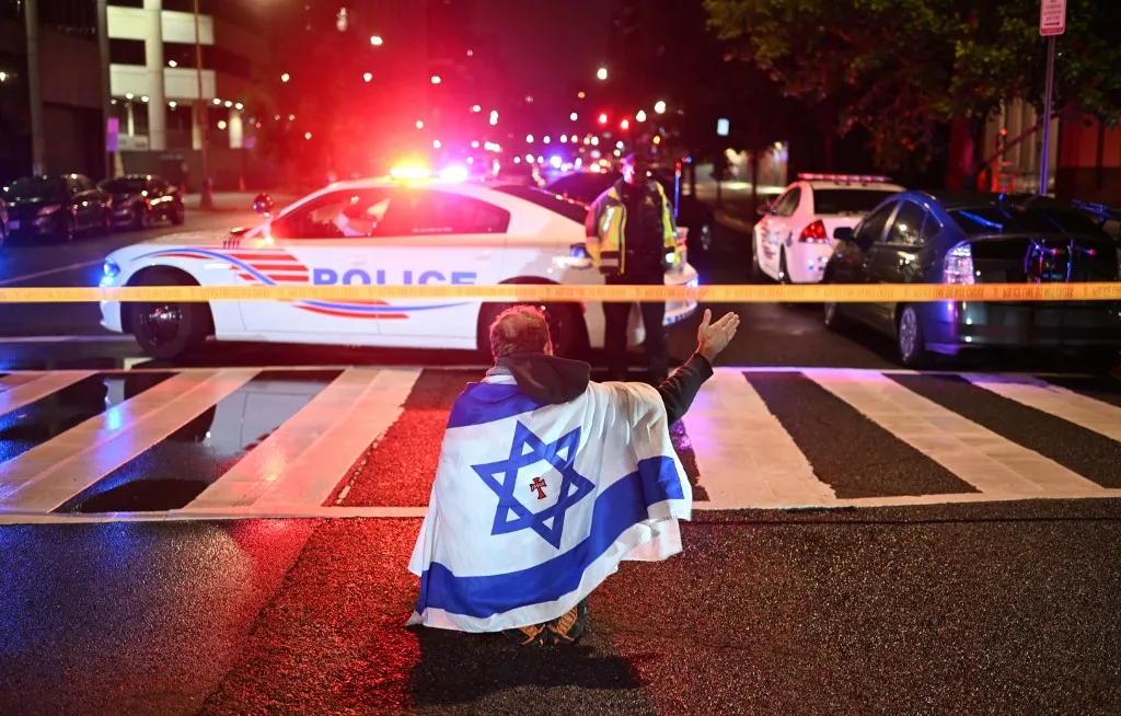 A man draped in the Israeli flag, bearing a cross and the name "Jesus" at its center, gestures as Metropolitan Police officers secure the area outside the Capital Jewish Museum following a shooting that left two people dead in Washington, DC, in the early hours of May 22, 2025. Two Israeli embassy staffers were shot dead late Wednesday, May 21, outside a Jewish museum in Washington by a gunman who shouted "free Palestine," authorities said, with US officials and Israeli diplomats expressing shock and outrage over the killings. (Photo by Alex WROBLEWSKI / AFP)
