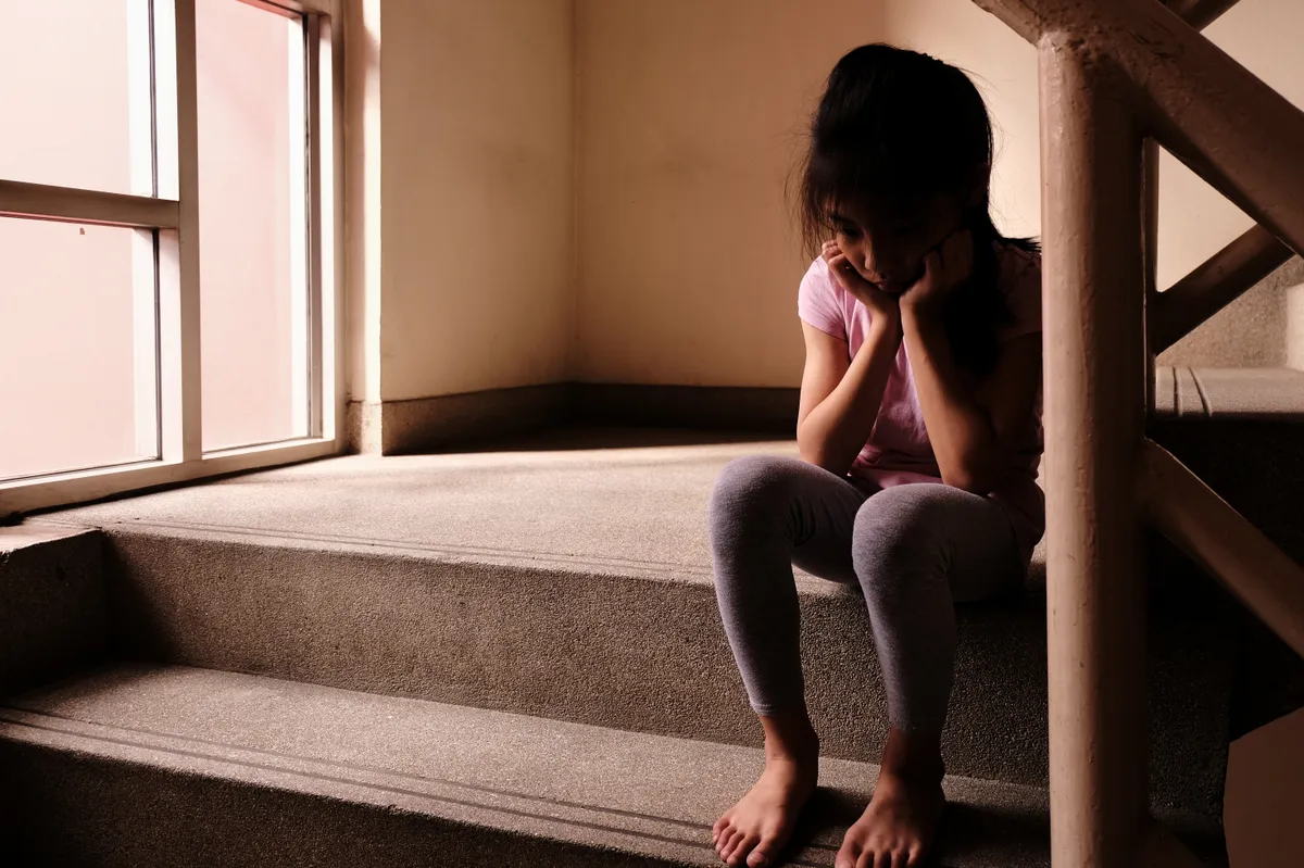 molesztálta, Child abuse concept. A sad and lonely young girl sitting by a stair inside a build with light through glass window in the background.