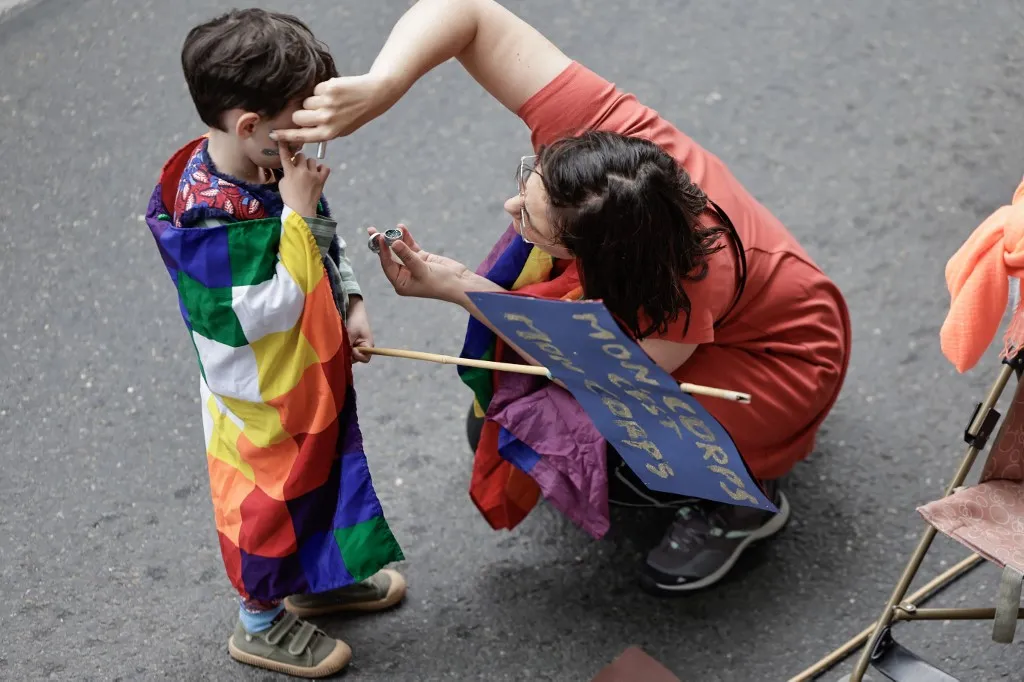A woman paints the face of a child wrapped in a rainbow flag holding a placard reading "My body is my body" before departing from La Courneuve - Aubervilliers RER station to take part in the "Suburbs Pride" at La Courneuve, a northern Paris suburb, on June 7, 2025. (Photo by STEPHANE DE SAKUTIN / AFP)