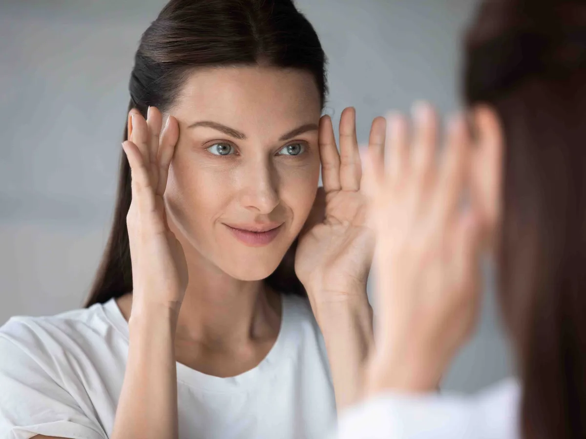 Smiling beautiful young woman look in mirror massage tap healthy glowing facial skin, pretty millennial female do morning beauty skincare face procedures or treatment, cosmetology concept