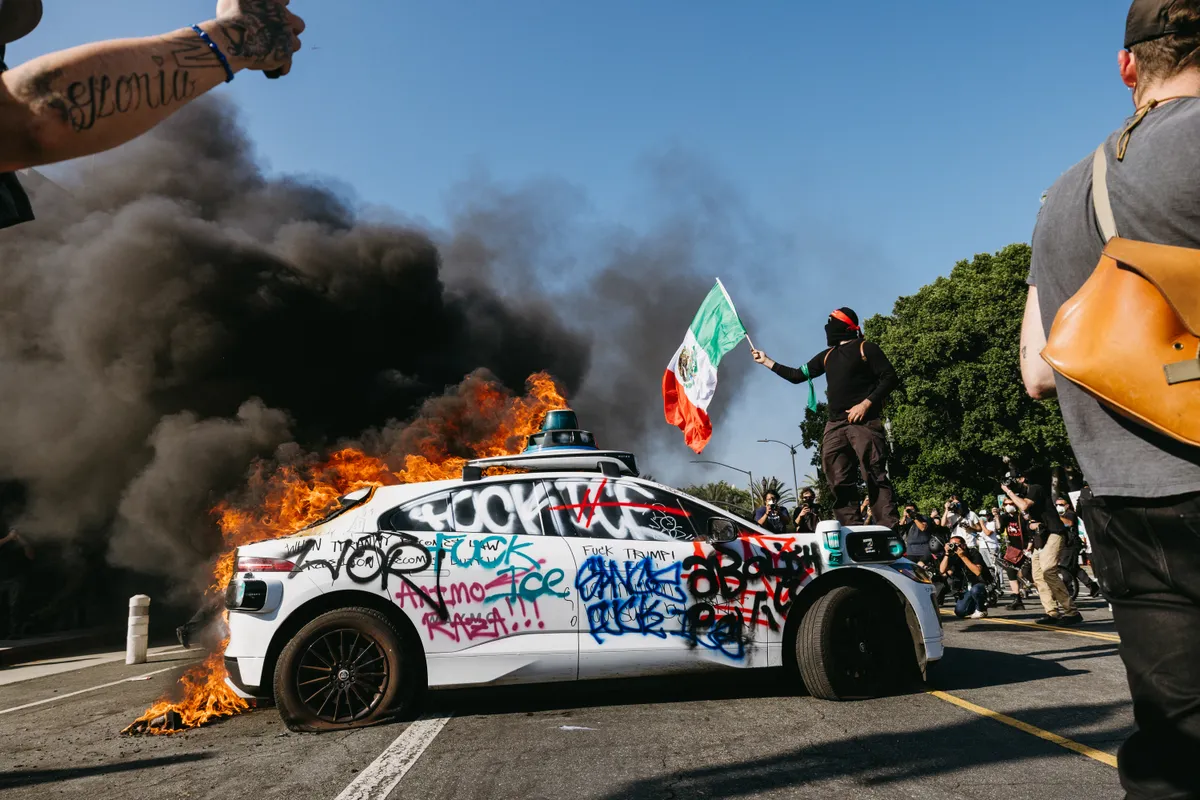 A Waymo vehicle burns in the street as smoke billows during protests in Los Angeles, California, on June 8, 2025. Following a series of aggressive federal immigration operations in Los Angeles, tensions escalated when Immigration and Customs Enforcement (ICE) agents conducted a high-profile raid on a Home Depot location. The raid sparked widespread protests across the city, where demonstrators decried the targeting of immigrant communities and the separation of families. Clashes soon broke out between protesters and federal agents, prompting U.S. President Donald Trump to deploy 2,000 National Guard troops to the city, a move harshly criticized by California officials, including the governor, as "purposefully inflammatory." (Photo by David Pashaee / Middle East Images via AFP), klubvilágbajnokság