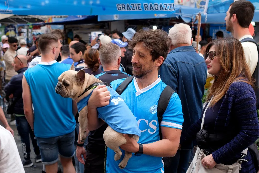 A man wearing a Napoli jersey holds a dog with a Napoli jersey as he celebrates the victory of the fourth Italian football Campions by Napoli in the Quartieri Spagnoli, downtown Naples, on May 24, 2025 after the match between Napoli and Cagliari held at the Diego Armando Maradona stadium on May 23, 2025. (Photo by Eliano Imperato / Controluce via AFP)