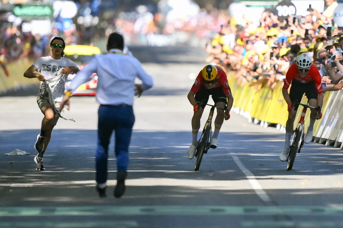 A Tour de France staff member attempts to intercept an individual wearing a t-shirt which reads "Israel out of the Tour" running at the finish line area as Uno-X Mobility team's Norwegian rider Jonas Abrahamsen (C) cycles ahead of Team Jayco AlUla team's Swiss rider Mauro Schmid (R) to the finish line to win the 11th stage of the 112th edition of the Tour de France cycling race, 156.8 km starting and finishing in Toulouse, southwestern France, on July 16, 2025. (Photo by Marco BERTORELLO / AFP)