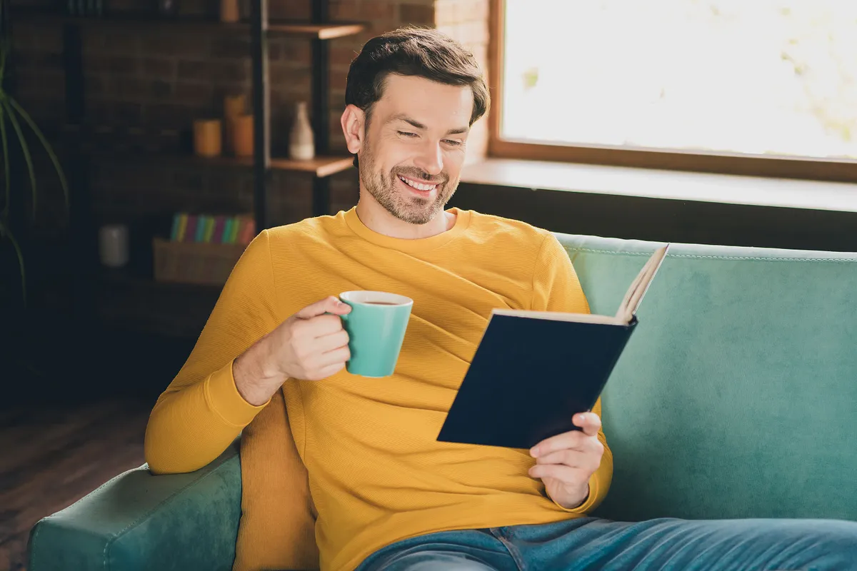 Photo portrait of handsome mature man read book drink tea sit couch dressed yellow outfit cozy day light home interior living room