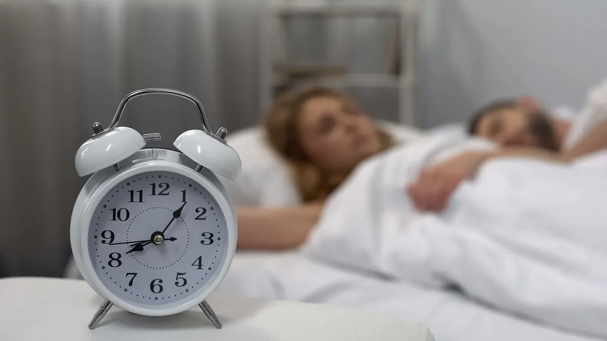 Az óraátállítás miatt rövidebb lesz az éjszaka Young couple sleeping in bed with ringing alarm clock on table, sleep biorhythms