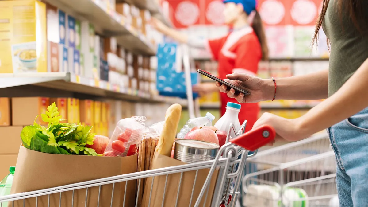 infláció, Woman buying groceries at the supermarket, she is pushing a shopping cart and using her smartphone, unrecognizable people