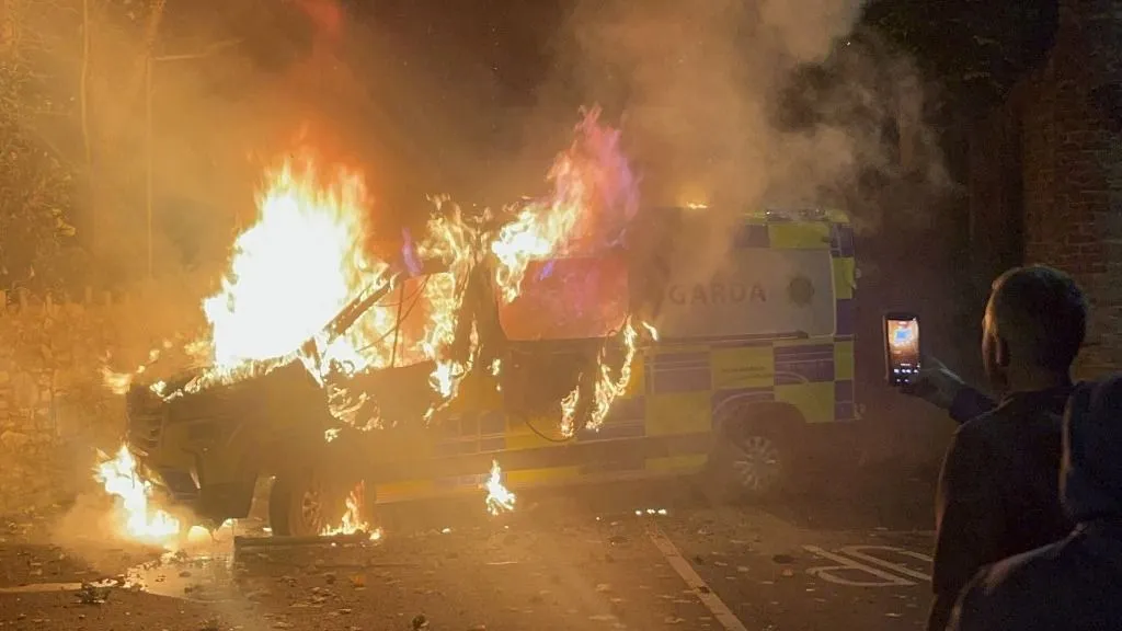 Onlookers film a police vehicle burning as a demonstration outside a hotel housing asylum seekers turns violent in Saggart, south-west of Dublin in Ireland on October 21, 2025. Police in riot gear clashed with at least 500 protesters in Dublin on Tuesday outside an asylum seeker hotel, following allegations that a 10-year-old girl was sexually assaulted. (Photo by Peter MURPHY / AFP)