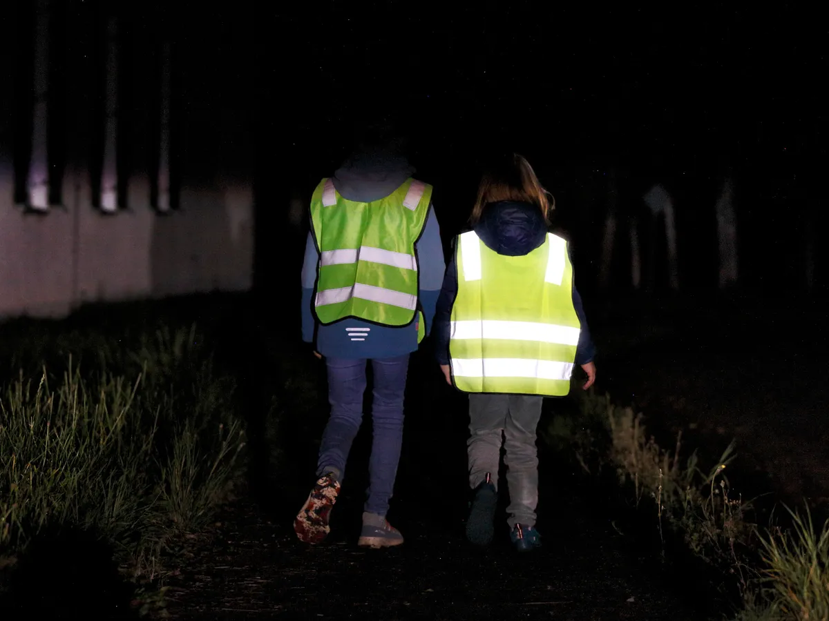 Egy nem reflektáló és egy jó láthatósági mellényt viselő gyermek áll egymás mellett a sötétben.
