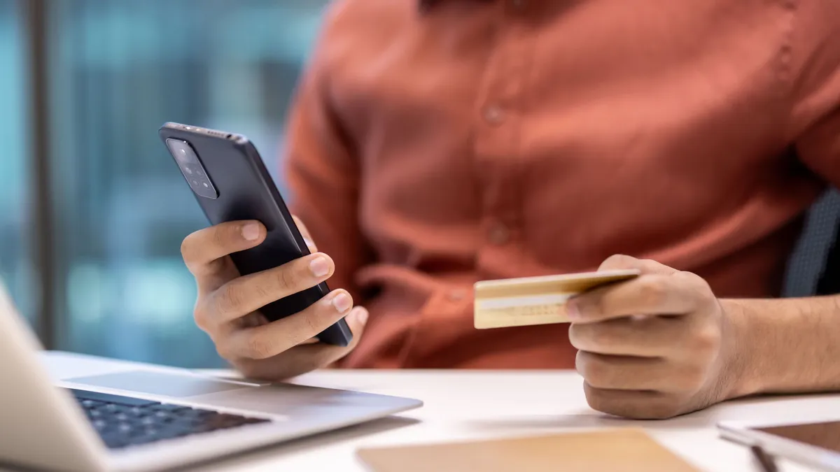  fix 3 százalékos hitel, Person holding phone and credit card while using laptop at office desk. Concept of online shopping, e-commerce, technology in business.