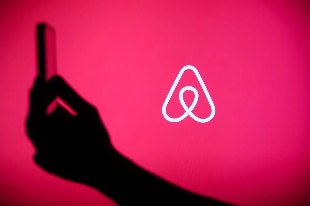 The Airbnb logo displays on a screen with a person holding a phone in Knurow, Poland, on September 20, 2025. (Photo by Klaudia Radecka/NurPhoto) (Photo by Klaudia Radecka / NurPhoto via AFP)