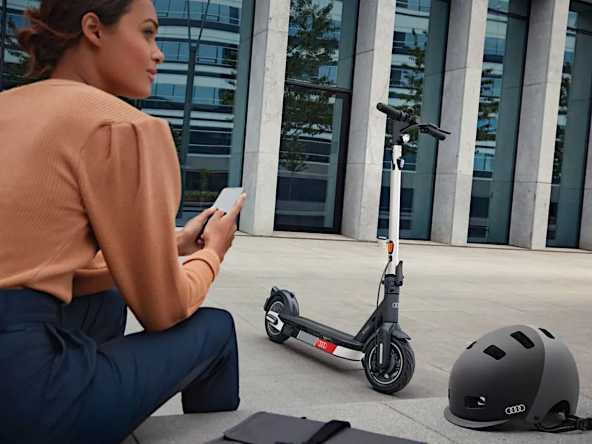 Egy e-roller parkol egy telefonját használó hölgy és annak bukósisakja között.