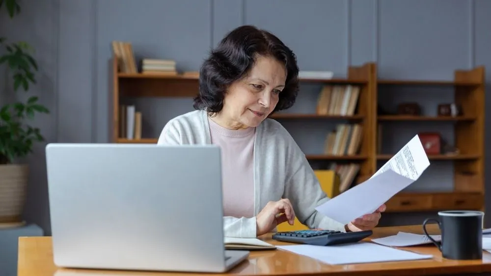 Middle aged senior woman sit with laptop and paper document. Pensive older mature lady reading paper bill pay online at home managing bank finances calculating taxes planning loan debt pension payment, NAV
