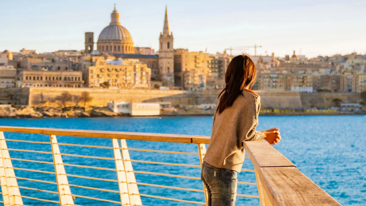 Málta, An Asian woman gazes at the stunning sunset over the ancient skyline of Valletta, Malta. The colorful balconies and vibrant architecture create a picturesque setting, perfect for relaxation.