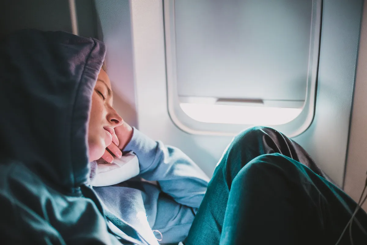 Tired blonde casual caucasian woman wearing sporty hoodie napping on seat while traveling by airplane. Commercial transportation by planes. Authentic image of real people
