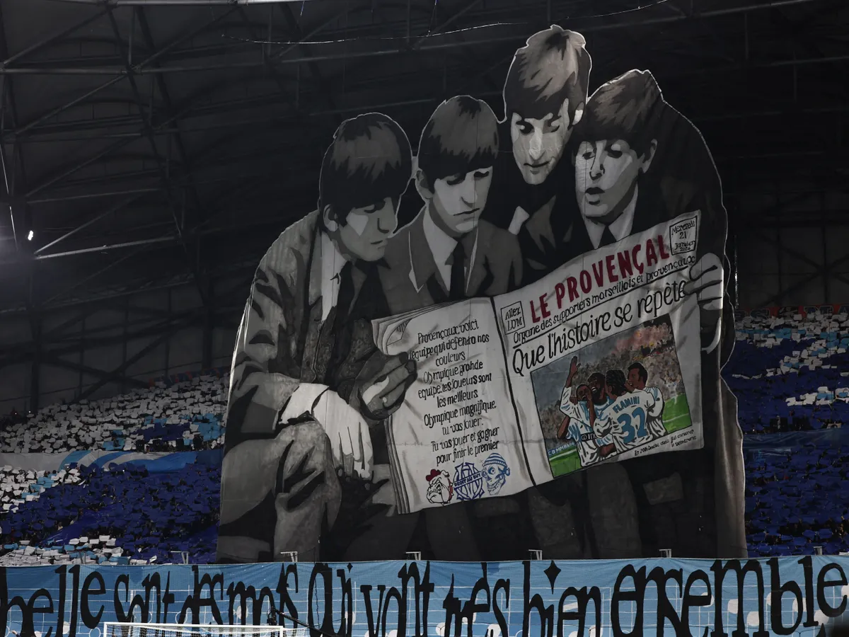 Supporters raise a banner in the likeness of members of the Beatles reading a copy of a southern French regional newspaper in the stands prior to the UEFA Champions League, league phase day 7, football match between Olympique de Marseille (OM) and Liverpool FC at the Stade Velodrome in Marseille, southern France, on January 21, 2026. (Photo by Thibaud MORITZ / AFP)