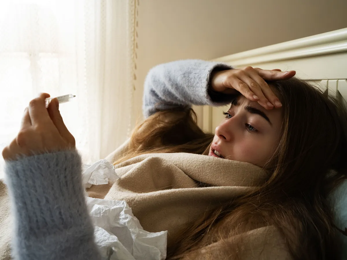 Woman check thermometer measure body temperature using glass thermometer hand on head lying in bed in the bedroom at home. Seasonal cold, flu epidemic, fever, sickness, illness, headache, influenza.