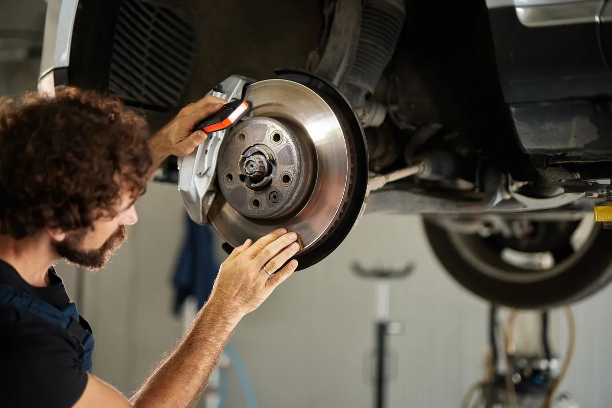 A skilled mechanic inspects and repairs the brake system of a car in a well-lit repair shop. Tools and equipment are organized around him, indicating an active operate session.