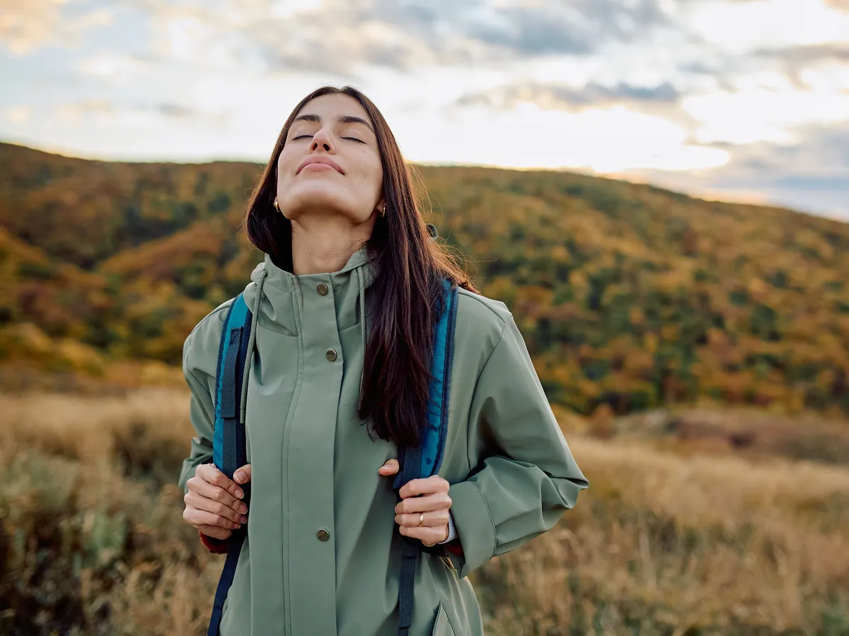 Woman standing on a mountain trail, eyes closed, breathing fresh air and feeling serene during an autumn hike