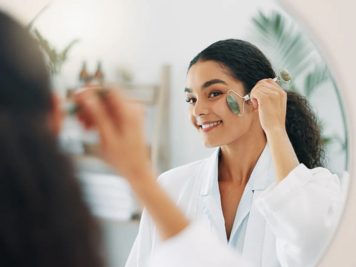 Mirror, happy woman and face roller for skincare or morning routine in home bathroom. Beauty, jade stone and reflection for facial cosmetic massage, treatment and lymphatic drainage for dermatology