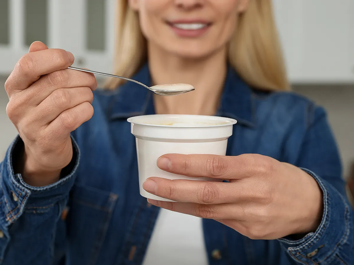 Woman holding plastic cup and spoon with tasty yogurt in kitchen, closeup