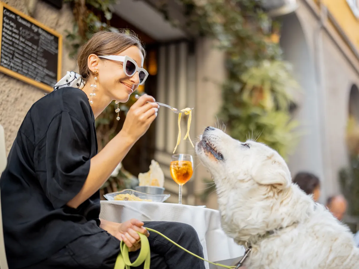 Woman eating pasta with her cute white dog at the restaurant outdoors. Concept of friendship with pets and having fun together