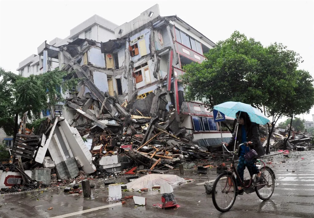 A cyclist rides past a dead body of an earthquake victim covered with plastic in front of a collapsed building in Dujiangyan southwest China Sichuan province on May 13, 2008. China was reeling May 13 from its worst earthquake in three decades which left tens of thousands of people dead, missing or trapped under crushed houses, schools and factories.  AFP PHOTO/TEH Eng Koon (Photo by TEH ENG KOON / AFP)