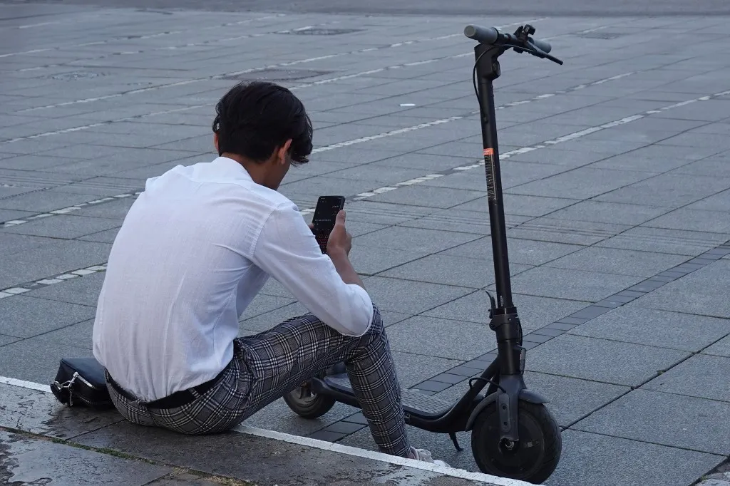 A young man wearing a white shirt and checked trousers sits on the edge of a pavement next to an electric scooter, looking at his smartphone in Stuttgart, Baden-Wuerttemberg, Germany, on June 20, 2025. (Photo by Michael Nguyen/NurPhoto) (Photo by Michael Nguyen / NurPhoto via AFP)