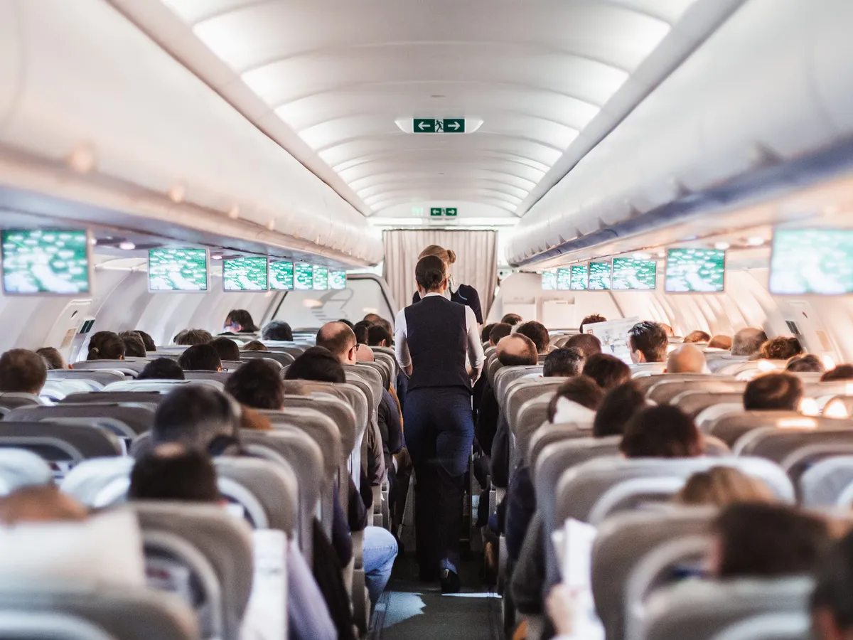 Interior of airplane with passengers on seats and stewardess in uniform walking the aisle, serving people. Commercial economy flight service concept