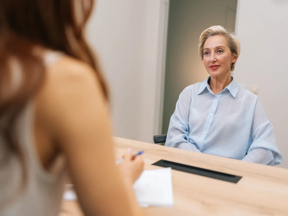 View from shoulder of 50s aged female job applicant sit at desk at employer office talk to recruiter answer questions on interview. Two business partners hold negotiations discuss deal, slow motion.