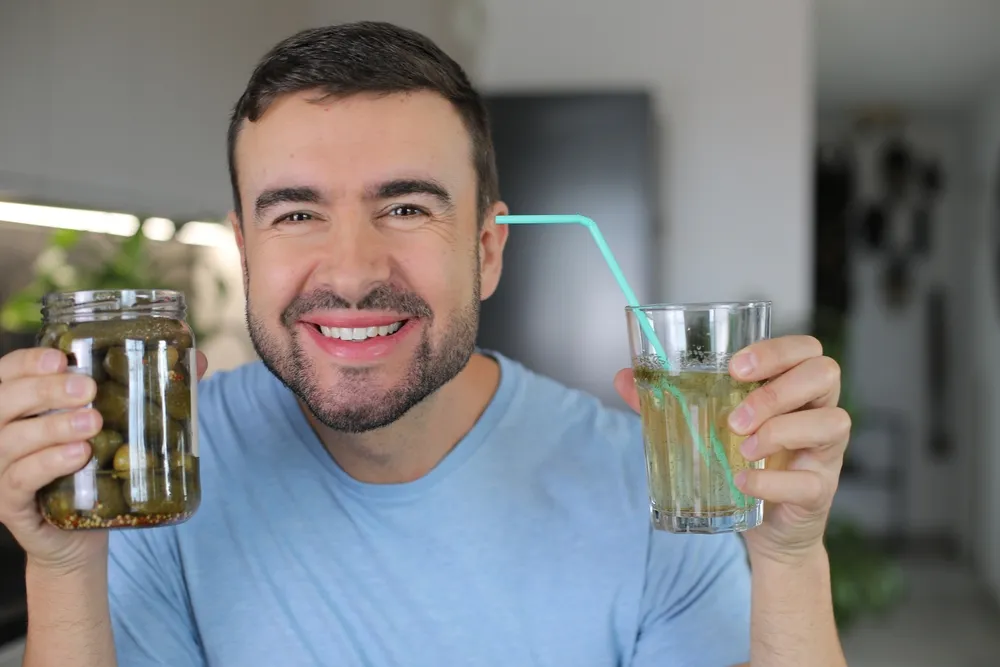 Egy üveg kovászos uborka és a kovászos uborka leve pohárban egy mosolygó férfi kezében