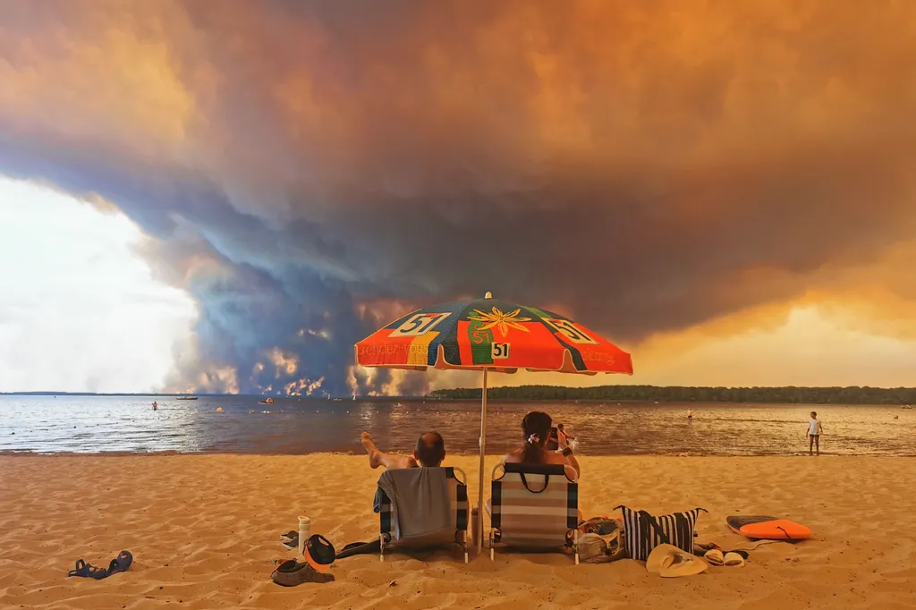 Beach goers rest on the beach of Moutchic on the shore of Lake Lacanau (Etang de Lacanau) in Lacanau , southwestern France on July 24, 2026, as clouds of smoke rise into the sky from the Gironde wildfire. The fire raging in the northern part of the Arcach