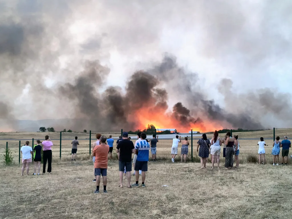 Bystanders look on as fire and smoke billow across Castres-Mazamet aerodrome, near Labruguiere in south-western France on July 24, 2026. France and Spain evacuated more than 160,000 people as unprecedented forest fires ravaged areas on July 25, 2026, near
