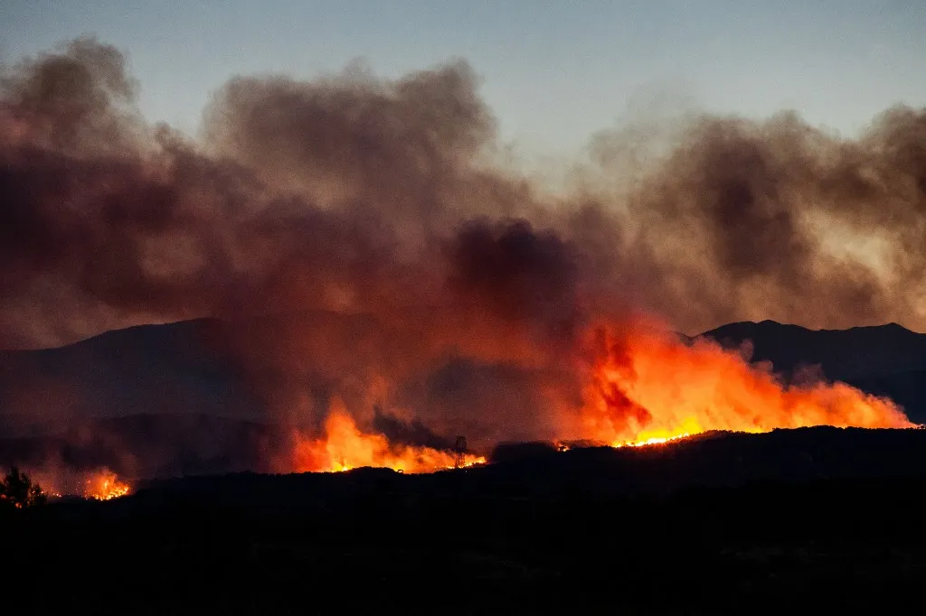 This photograph shows a wildfire burning in the Aspres region seen from Millas, in the Pyrenees-Orientales department, southern France on July 5, 2026. A wildfire broke out on July 4 at around 19h30 (1730 GMT) in a hard-to-reach massif near Trevillach, in the Pyrenees-Orientales department of southern France and spread rapidly overnight. More than 10,000 residents are currently being evacuated as the wildfire spreads rapidly, having burned around 2,000 hectares. (Photo by Jc Milhet / AFP)