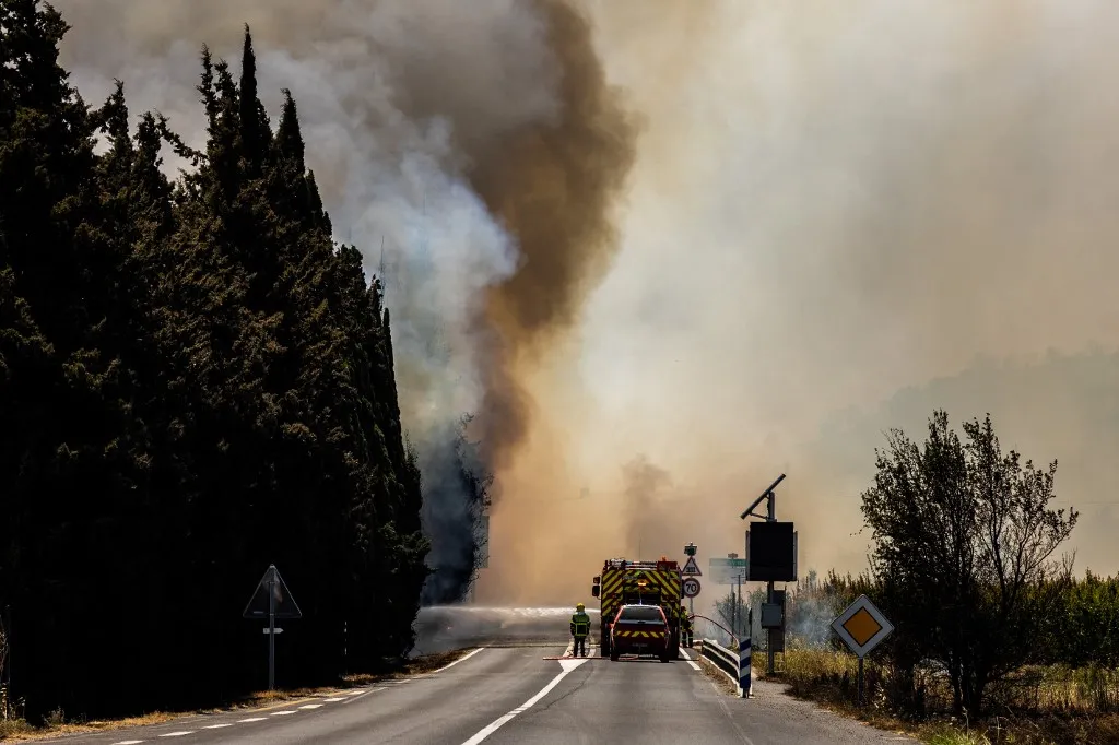 A firefighter sprays water on flames as a wildfire rages in the southern France department of Pyrenees-Orientales near the town of Bouleternere, near Perpignan on July 6, 2026. 700 firefighters backed by special aircraft battled to control a "gigantic" blaze spreading in a hard-to-reach remote area near Ille-sur-Tet. Wildfires raged across southern Europe, forcing thousands of people to evacuate their homes and prompting officials to ban spectators from a stage of the storied Tour de France cycling race. (Photo by Jean-Christophe MILHET / AFP)