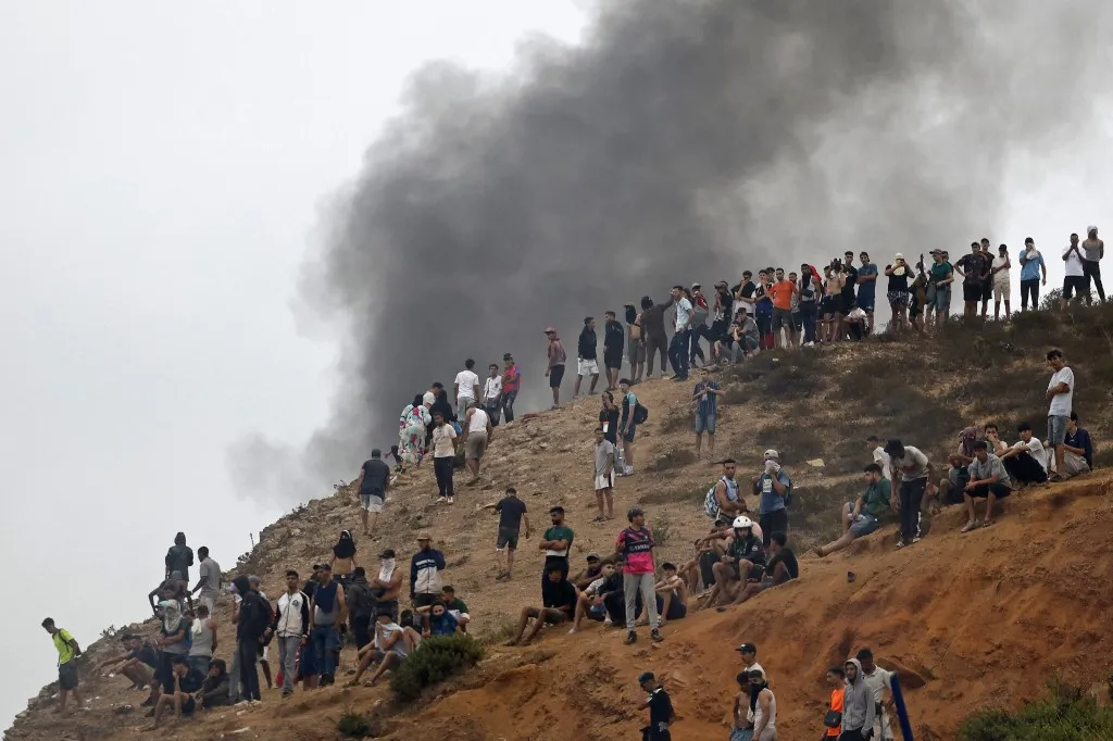 People gather on a hilltop as smoke billows from the site of clashes near Fnideq, on the Morocco-Spain border, on July 31, 2026. Thousands of migrants crossed overnight from Morocco into Spain's north Africa's Ceuta enclave, a police source said on July 31, adding that it was impossible to estimate how many people in total. A total of 18 people died while attempting to reach the Spanish enclave, the source said. (Photo by Abdel Majid BZIOUAT / AFP), migránsok, Ceuta, Spanyolország