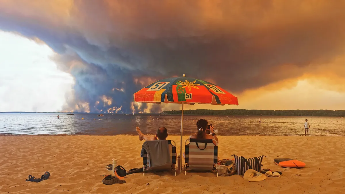 (FILES) Beach goers sit on the beach of Moutchic in Lacanau, southwestern France on July 24, 2026, as clouds of smoke rise into the sky from a wildfire. France has been battered by three heatwaves since May as part of increasingly common extreme weather episodes linked to climate change, causing thousands of excess deaths and fuelling wildfires. (Photo by Maximilien LAMY / AFP)