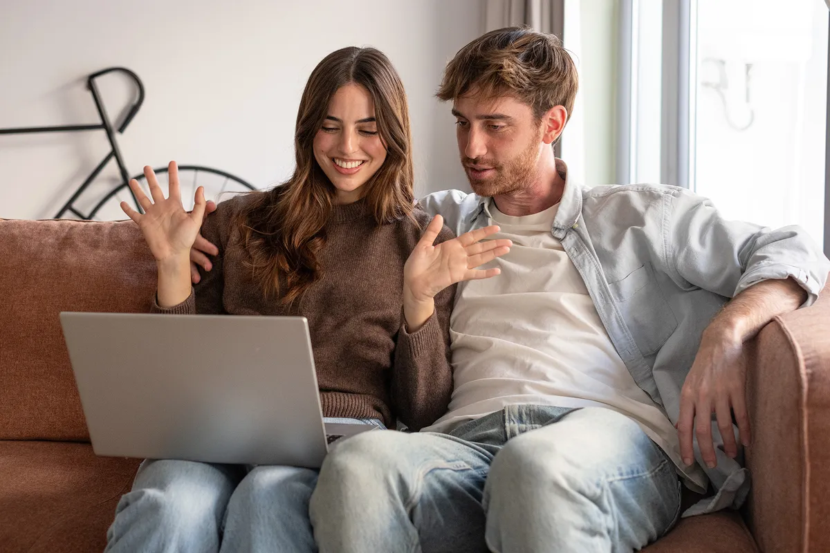 A happy young couple sits on the couch, staring at the computer screen, gesturing and looking overjoyed. They're screaming with joy, reading fantastic news, receiving a great business offer, chatting in video call