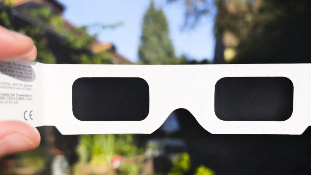 napfogyatkozás, Close-up view of white CE marked solar eclipse glasses held in front of a sunlit garden and a house before the August 12, 2026 solar eclipse in Saint-Cosme-en-Vairais, Sarthe, France on August 10, 2026. The cardboard glasses are intended for direct solar viewing and illustrate the need for certified eye protection as protective eclipse glasses are becoming difficult to find before the event. (Photo by Jimmy Beunardeau / Hans Lucas via AFP)