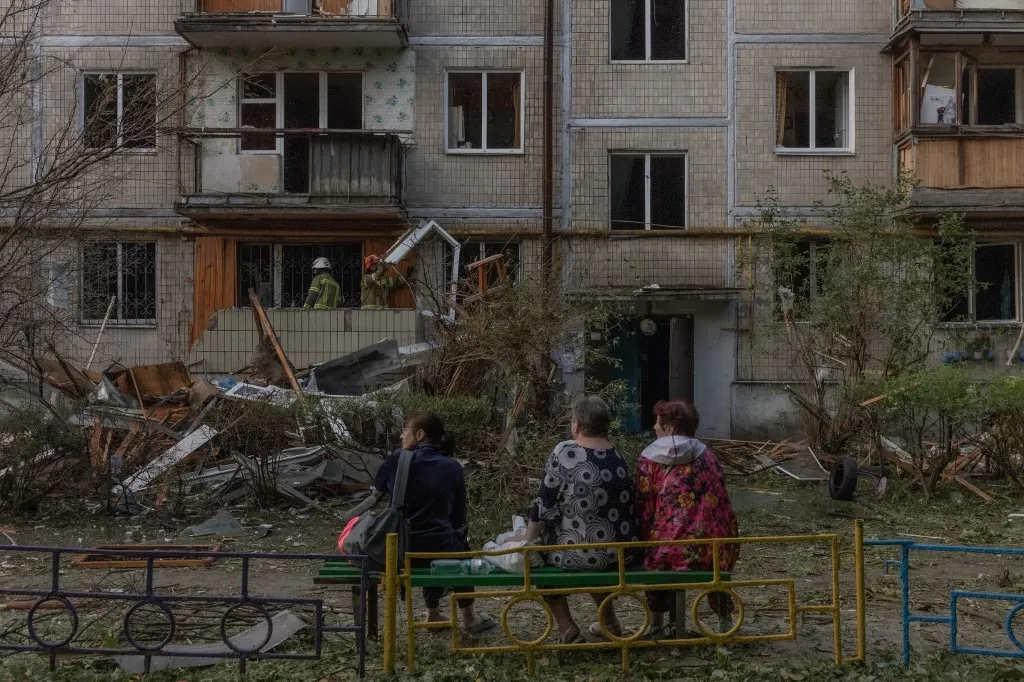Residents sit as rescuers work in a multi-storey residential building damaged in a Russian air attack on Kyiv on August 1, 2026, amid the Russian invasion of Ukraine. Russian strikes on Ukraine's capital killed at least nine people and wounded more than two dozen others, local officials said, hours after Kyiv's mayor had warned of a ballistic missile attack. (Photo by Roman PILIPEY / AFP), Ukrajna, Kijev, orosz rakétatámadás