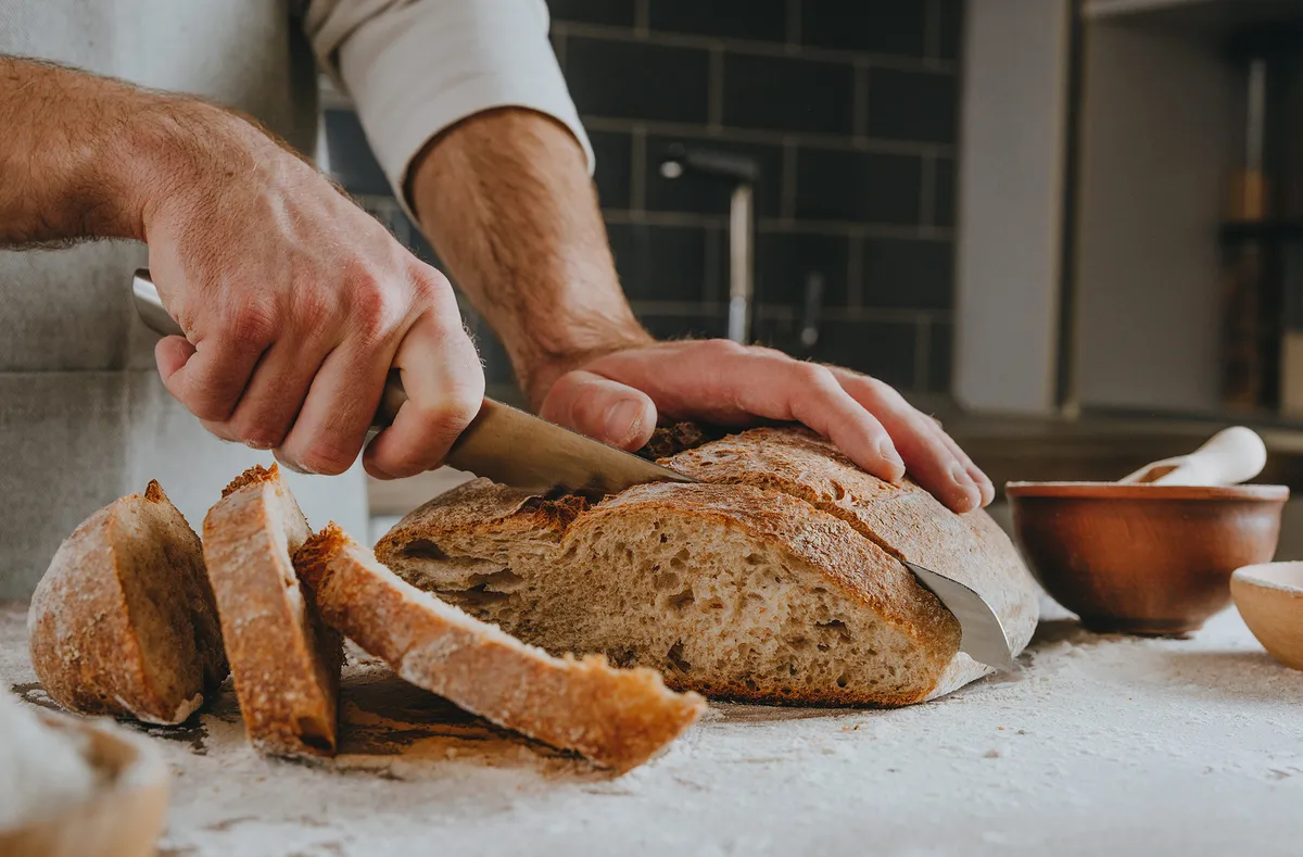 Unrecognizable young man in apron cutting homemade whole grain bread. selective focus.